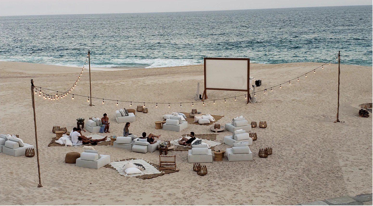 Outdoor movie screen and lounge seating on the beach under string lights